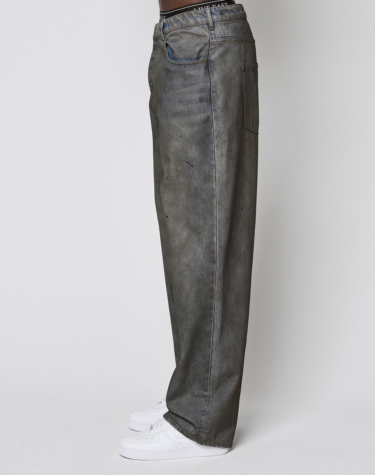 Model in LFDY Coated Baggy Denim by LIVE FAST DIE YOUNG: washed gray, baggy fit with wide straight leg, styled with white sneakers, shown from the side against a plain white background.