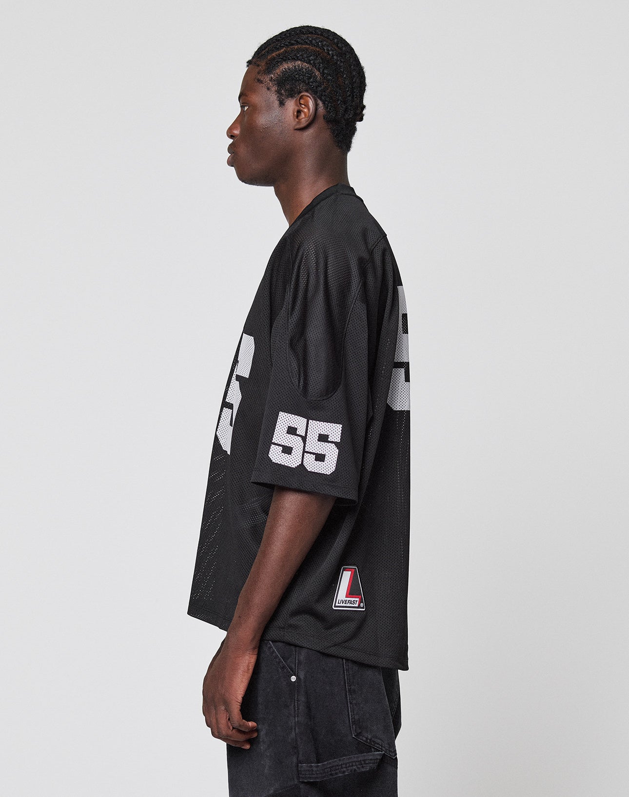A person with braided hair stands in profile wearing the LIVE FAST DIE YOUNG LFDY Mesh Jersey—an oversized black sports jersey featuring the number 55 in white on the sleeve and side—with dark pants, against a plain light grey background.