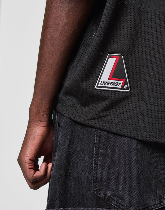 Close-up of a person's left arm and torso wearing the LFDY Mesh Jersey by LIVE FAST DIE YOUNG, featuring an oversize fit, black mesh fabric, and a striking red, white, and black patch. Styled with black denim shorts for a relaxed, on-trend look.