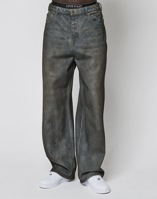 A person wearing LIVE FAST DIE YOUNGs LFDY Coated Baggy Denim—baggy fit, faded gray with black spots—and a black LIVE FAST waistband stands against a plain white background in white sneakers.