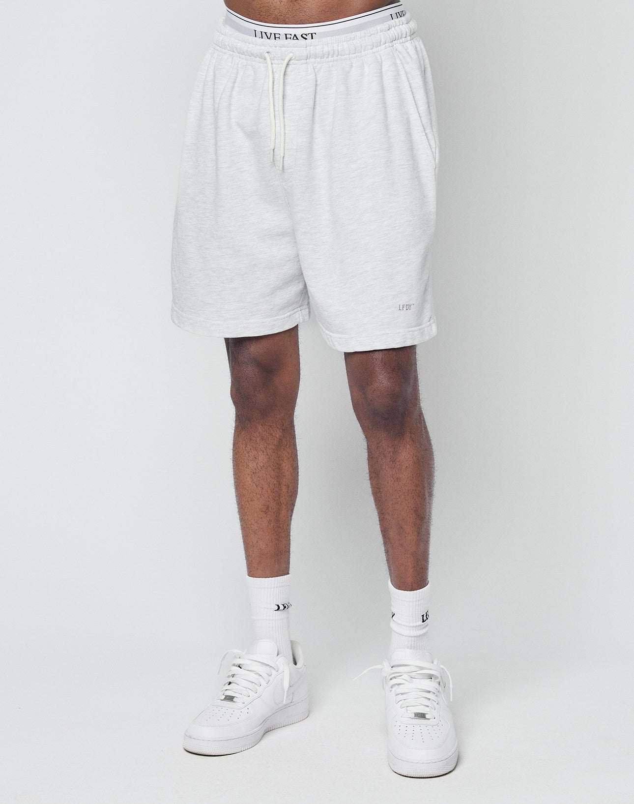 Someone wears LIVE FAST DIE YOUNG Basic Sweat Shorts in Regular Fit light gray with elastic waistband and visible logo print, styled with white crew socks and sneakers, standing against a plain light background.