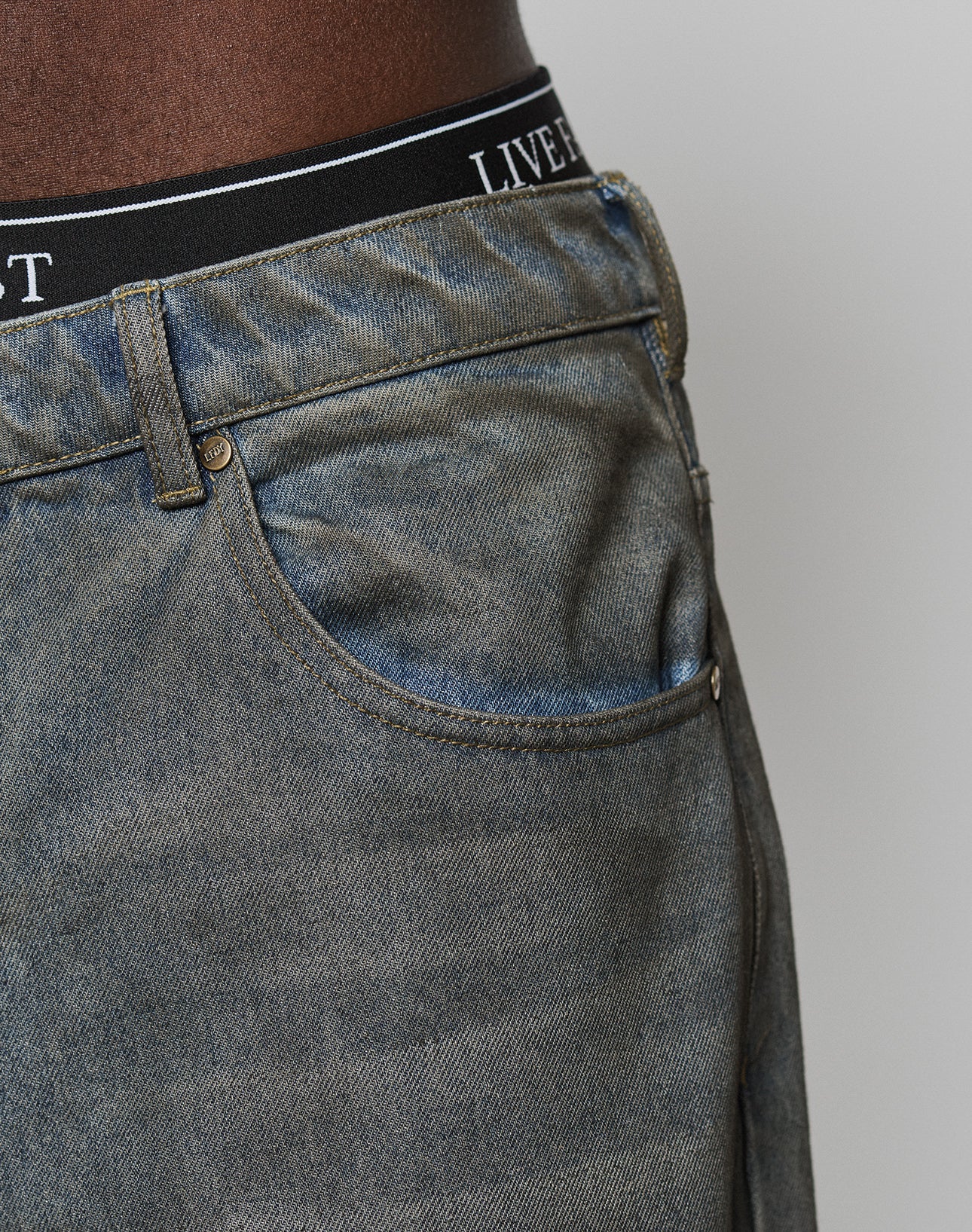 Close-up of a person wearing LFDY Coated Baggy Denim by LIVE FAST DIE YOUNG in faded blue, showing the front pocket and waistband details, paired with black underwear featuring LIVE on the waistband.