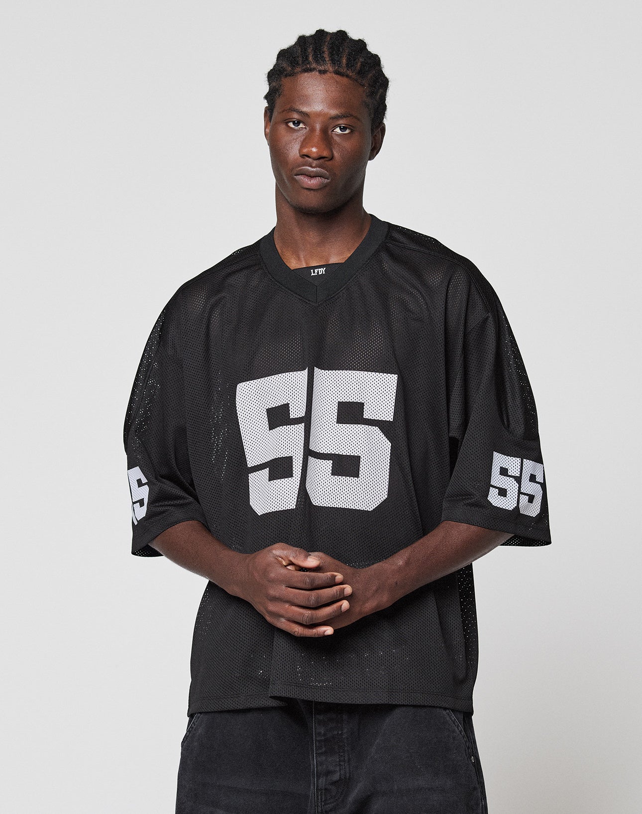 A man with braided hair stands against a plain background, wearing the oversize LIVE FAST DIE YOUNG LFDY Mesh Jersey with a V-neck and white number 55. His hands are clasped in front, highlighting the jersey’s relaxed fit.