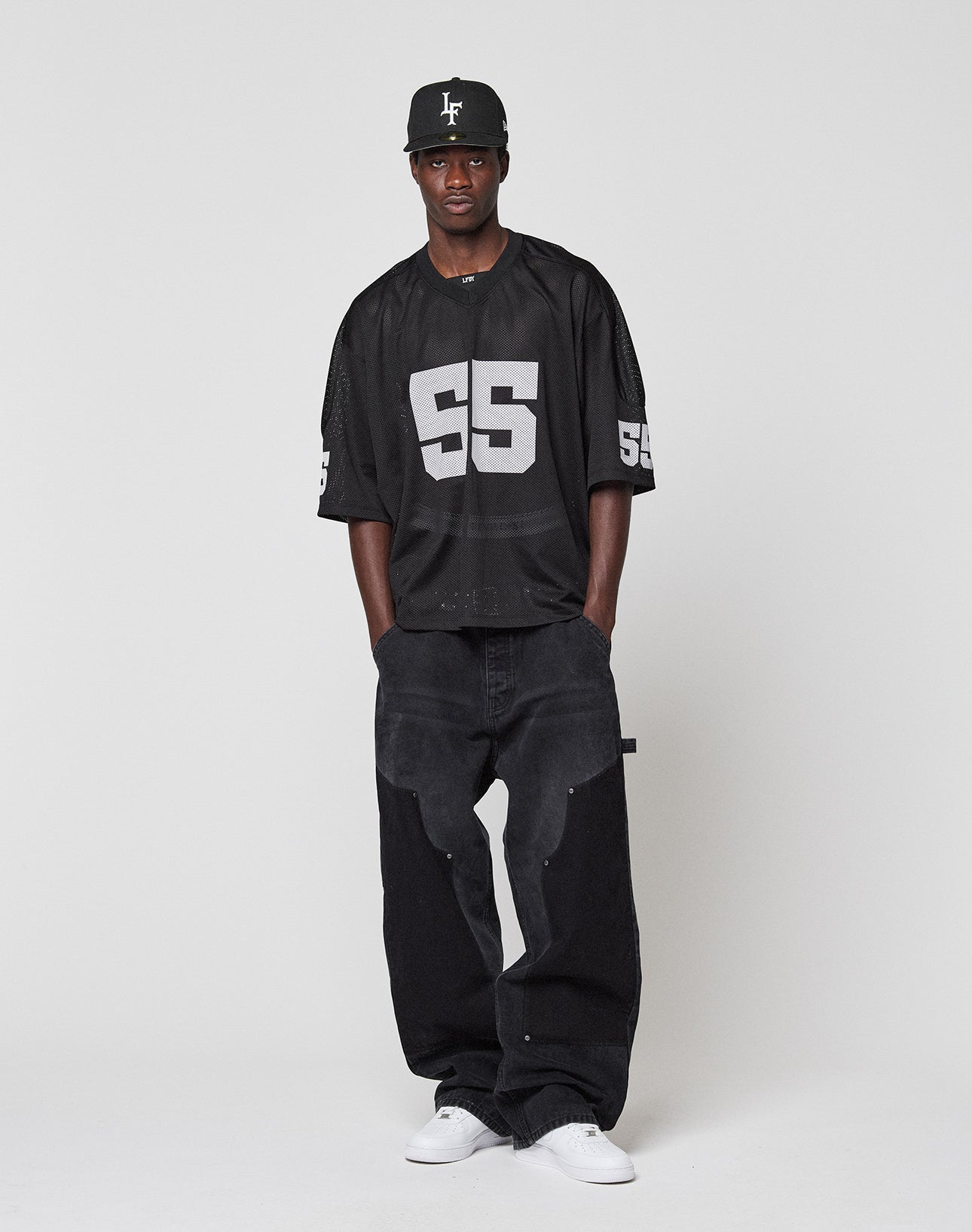 A man stands against a plain white background, wearing a black oversize baseball cap, baggy black jeans, white sneakers, and the LIVE FAST DIE YOUNG LFDY Mesh Jersey—a loose black polyester top with number 55 and V-neck.