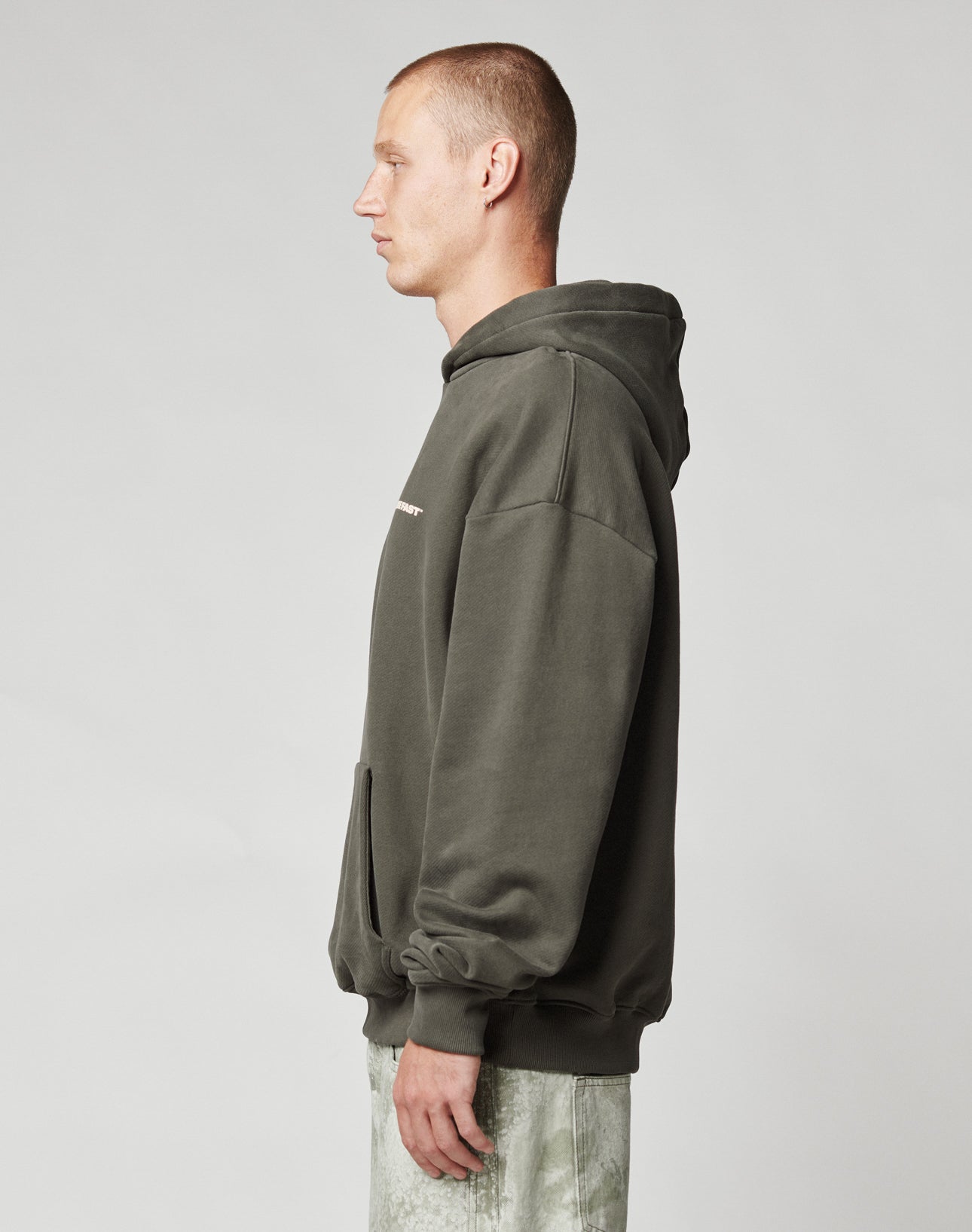 A person with short hair in profile wears the LIVE FAST DIE YOUNG Live Fast Plain Hooded, an oversize dark green soft Baumwolle hoodie with a front pocket, and light-colored pants, standing against a plain light gray background.
