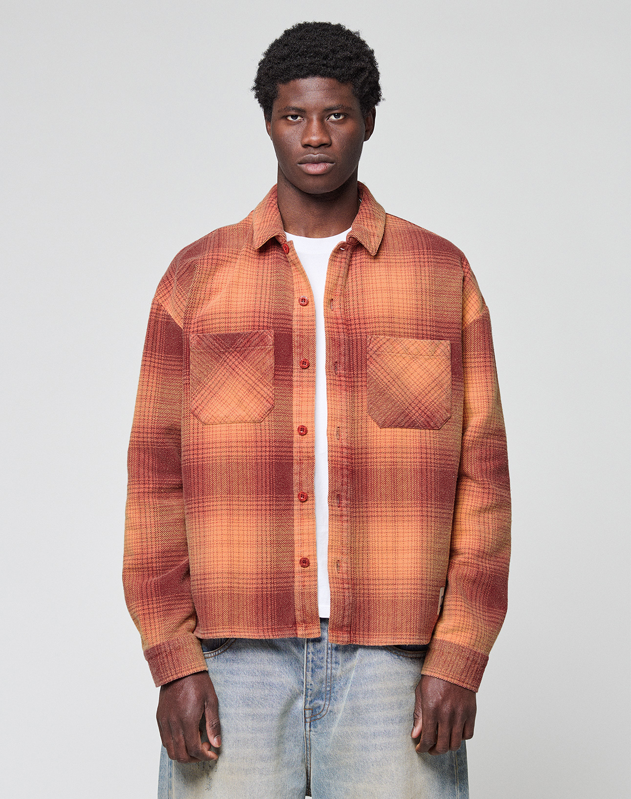 A person faces forward in a LIVE FAST DIE YOUNG Vintage Flannel (red and orange plaid) over a white tee and light blue vintage-wash jeans, standing against a plain light gray background.