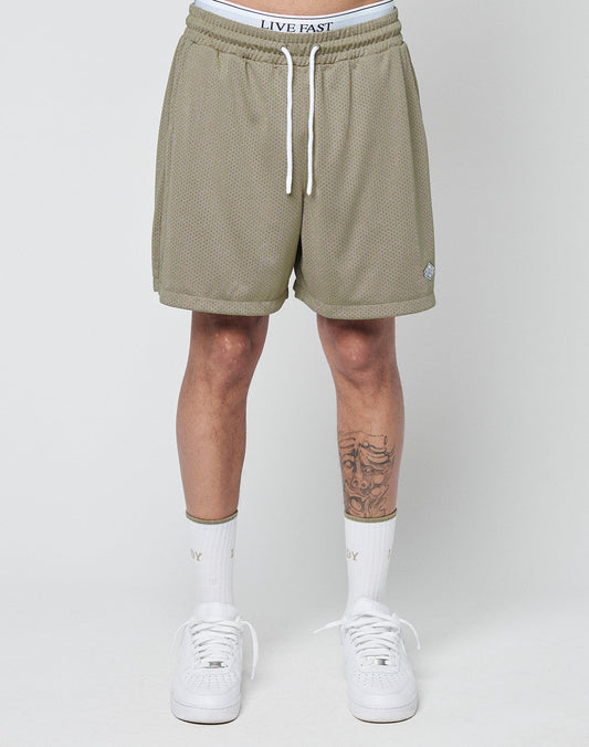 A person stands against a plain background wearing LIVE FAST DIE YOUNG Basic Mesh Shorts in olive green with an elastic waistband and white branded band, paired with white socks and sneakers; a tattoo is visible on their left shin.