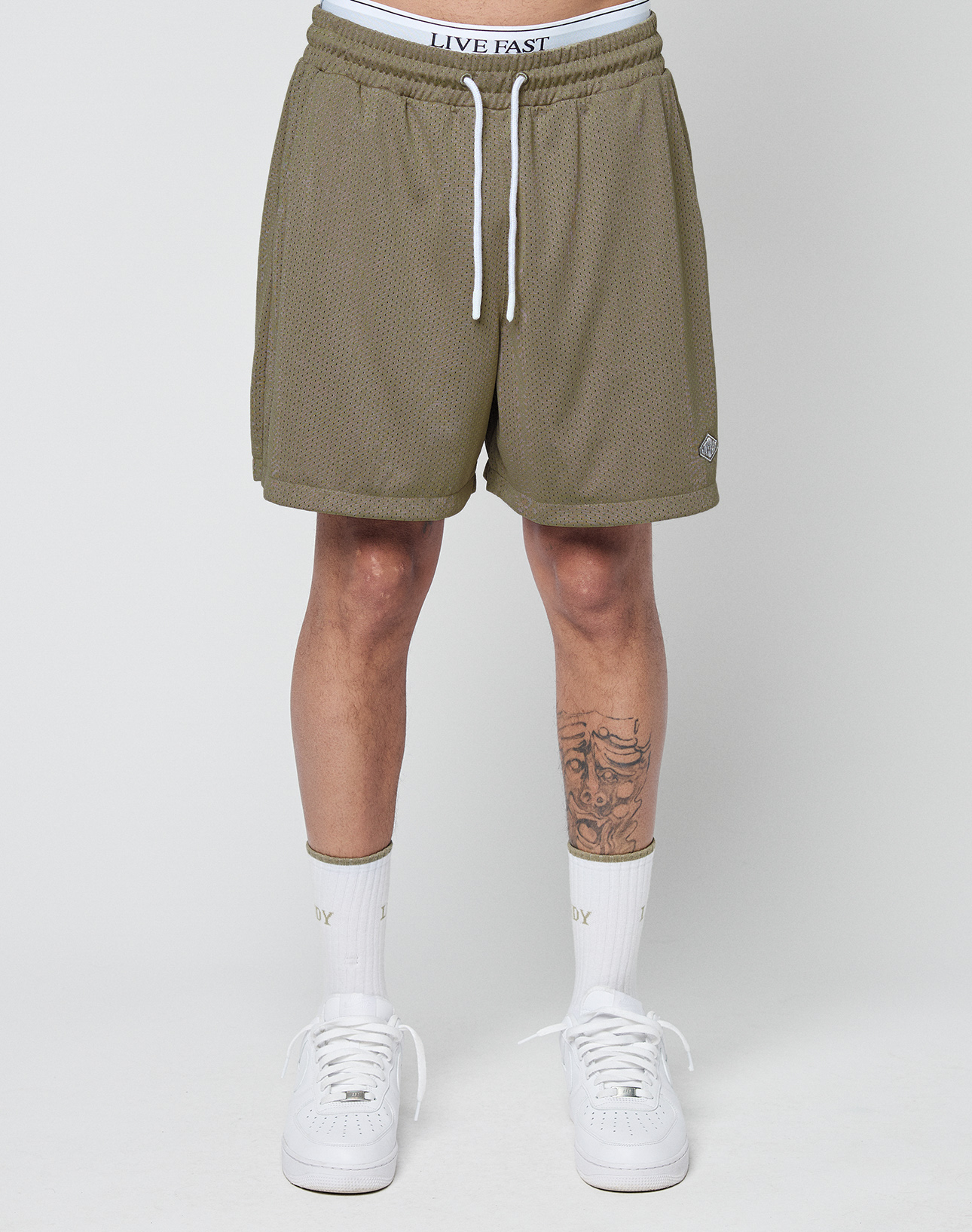 A person wears olive green LIVE FAST DIE YOUNG Basic Mesh Shorts with an elastic waistband, white LIVE FAST underwear, white socks, and sneakers. Only the lower half is shown; one leg has a tattoo. The background is plain and light-colored.