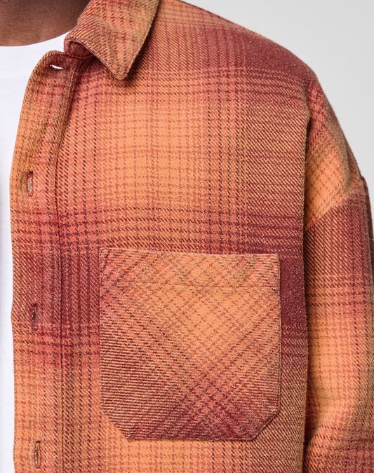 Close-up of a person wearing the LIVE FAST DIE YOUNG Vintage Flannel in plaid orange, brown, and yellow with vintage wash. The oversized shirt has a front pocket, visible buttons, and is layered over a white T-shirt.