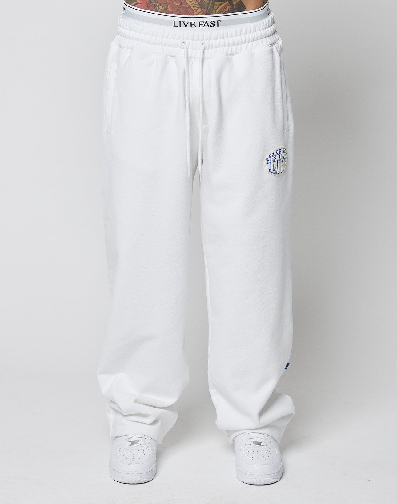 A person wears LFDY Radiator Sweatpants by LIVE FAST DIE YOUNG—oversized, white with elastic waistband and a small blue embroidered logo on the left thigh—paired with white sneakers. Tattoos show above the waistband against a plain white background.