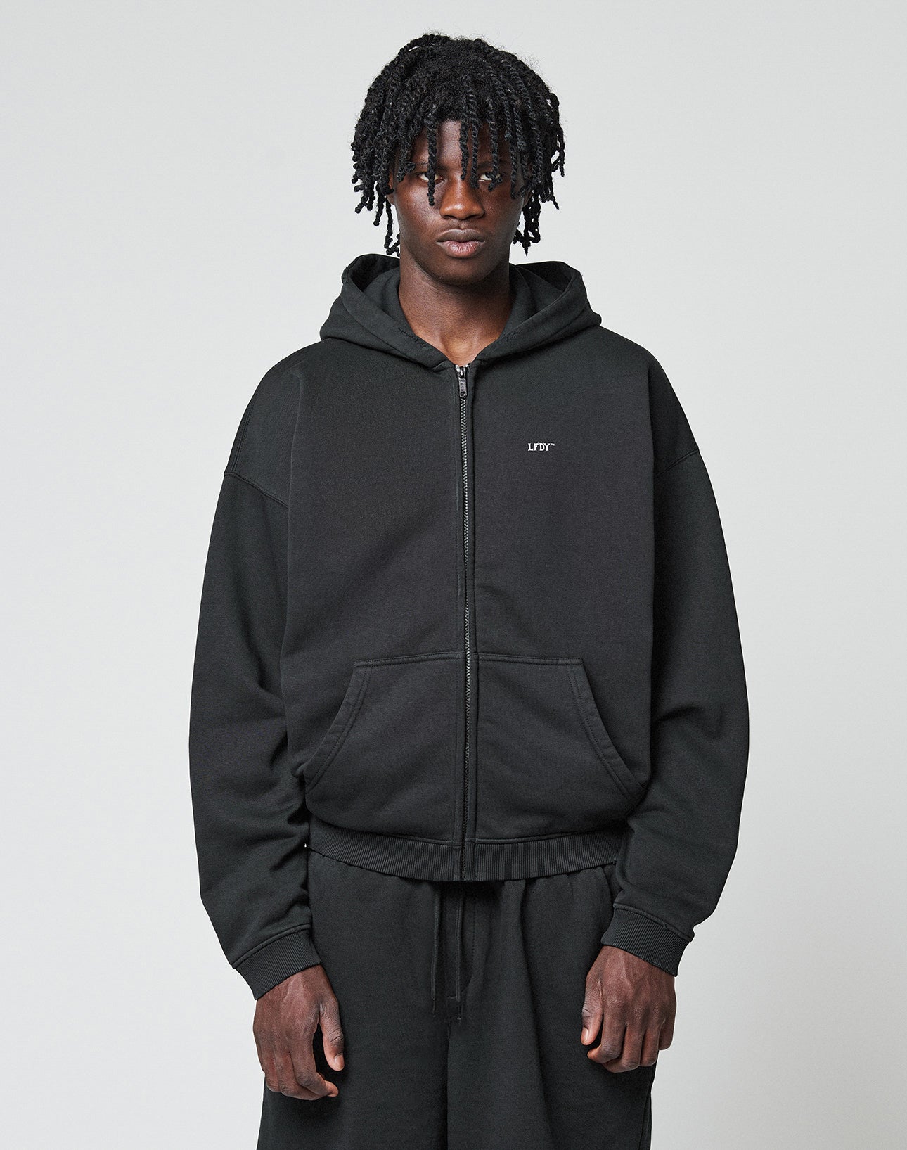 A person with medium-length twists wears a black LIVE FAST DIE YOUNG Basic Hooded Zip and black pants, standing against a plain light gray background, looking directly at the camera with a neutral expression. | model