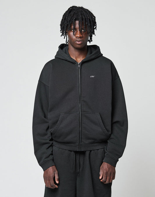 A person with medium-length twists wears a black LIVE FAST DIE YOUNG Basic Hooded Zip and black pants, standing against a plain light gray background, looking directly at the camera with a neutral expression. | model