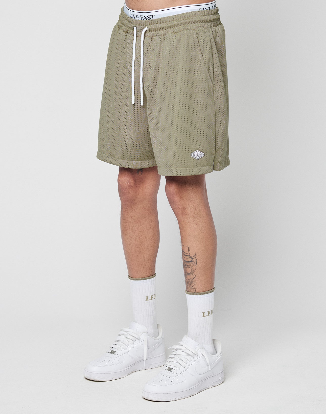 A person’s lower body is shown wearing LIVE FAST DIE YOUNG Basic Mesh Shorts in olive green polyester mesh, with an elastic waistband, drawstring, and a logo patch near the hem. They also wear white “LIVE FAST” socks and white sneakers.