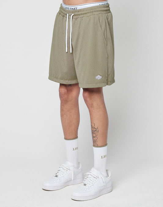 A person’s lower body is shown wearing LIVE FAST DIE YOUNG Basic Mesh Shorts in olive green polyester mesh, with an elastic waistband, drawstring, and a logo patch near the hem. They also wear white “LIVE FAST” socks and white sneakers.