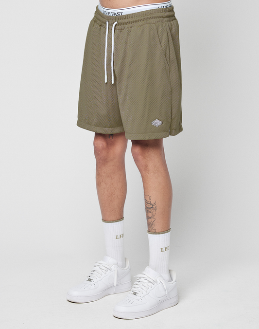 Wearing LIVE FAST DIE YOUNG Basic Mesh Shorts in olive green with an elastic waistband and logo patch, paired with a white LIVE FAST waistband, white socks, and sneakers. Only the lower half is shown against a plain white background. | model