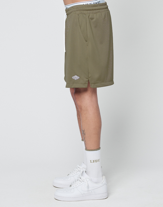 A person wearing olive green LIVE FAST DIE YOUNG Basic Mesh Shorts with a logo patch and elastic waistband, white crew socks with LFDY text, and white Nike sneakers stands sideways against a plain white background.