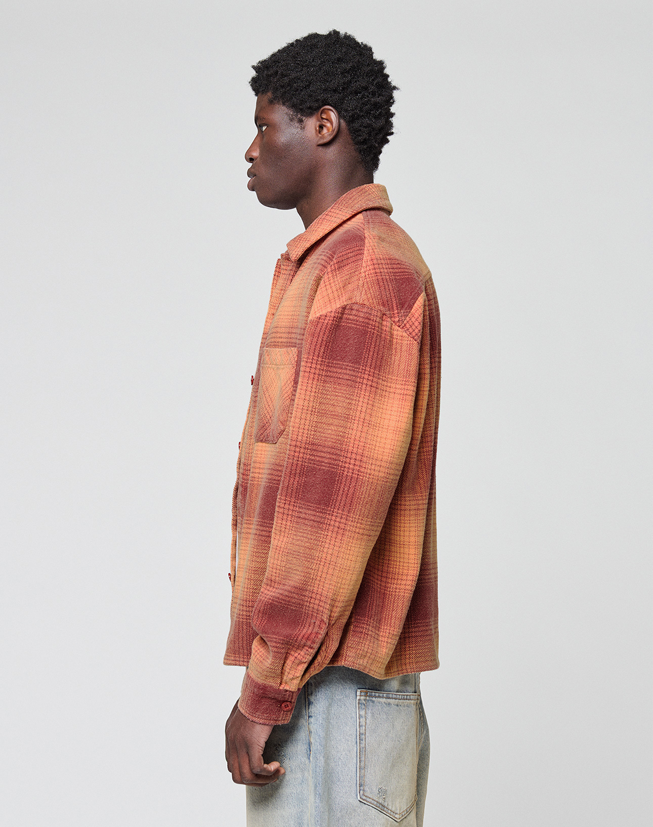 A person stands in profile against a plain background, wearing an oversize fit LIVE FAST DIE YOUNG Vintage Flannel with a red and orange plaid pattern and light blue jeans in vintage wash.