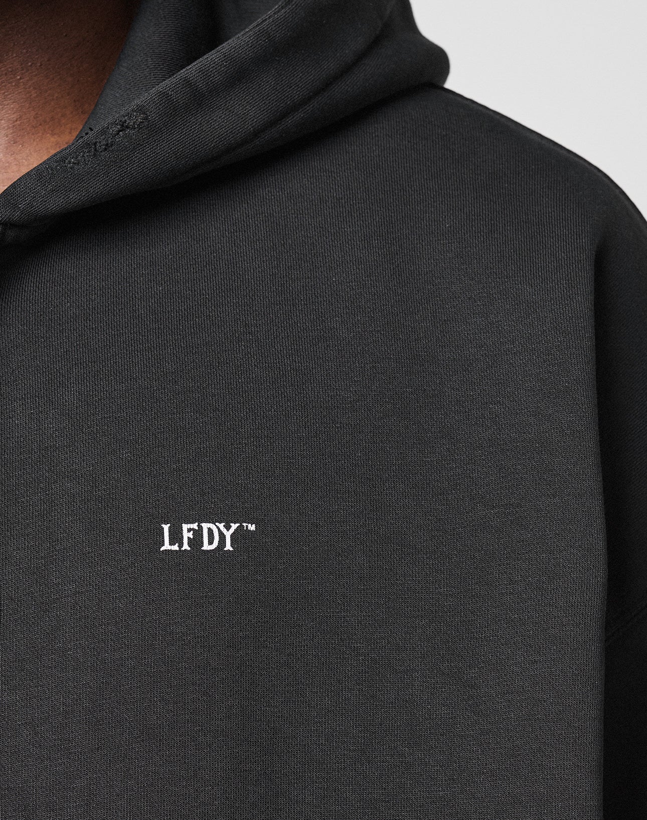 Close-up of a black LIVE FAST DIE YOUNG Basic Hooded Zip with hood up, featuring small white embroidered LFDY™ text on the chest and subtle distressed details. Only part of the wearer’s face and shoulder are visible.