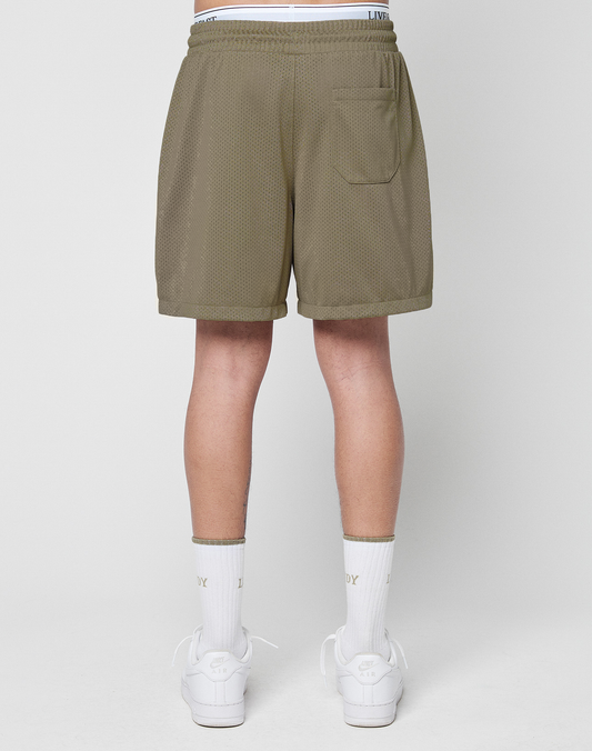A person faces away, wearing olive green LIVE FAST DIE YOUNG Basic Mesh Shorts with elastic waistband, white socks, and white sneakers. The shorts have a back right pocket and a subtle logo patch. Background is plain white.