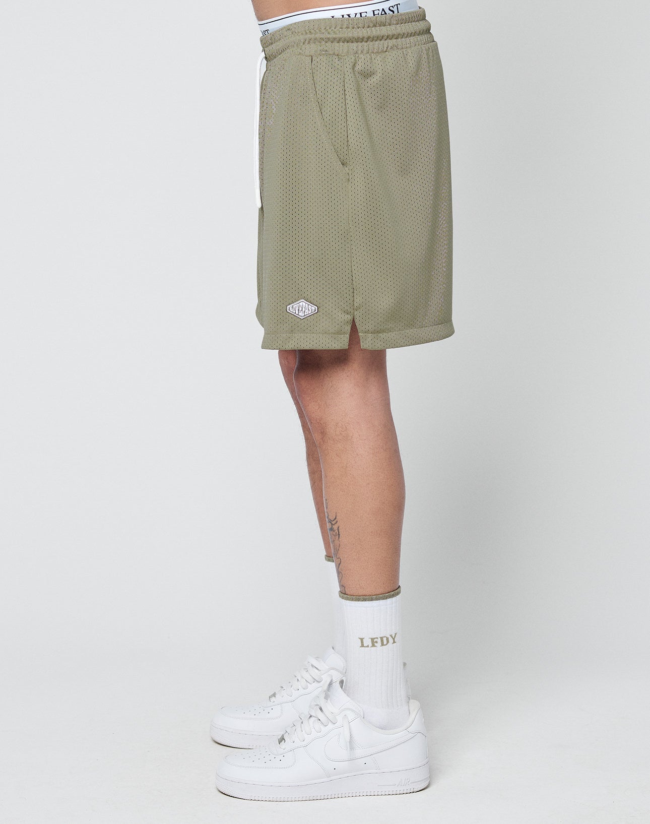 A person stands sideways in LIVE FAST DIE YOUNG Basic Mesh Shorts (olive green, logo patch, elastic waistband), white LFDY socks, and white Nike Air Force 1 sneakers against a plain light background.