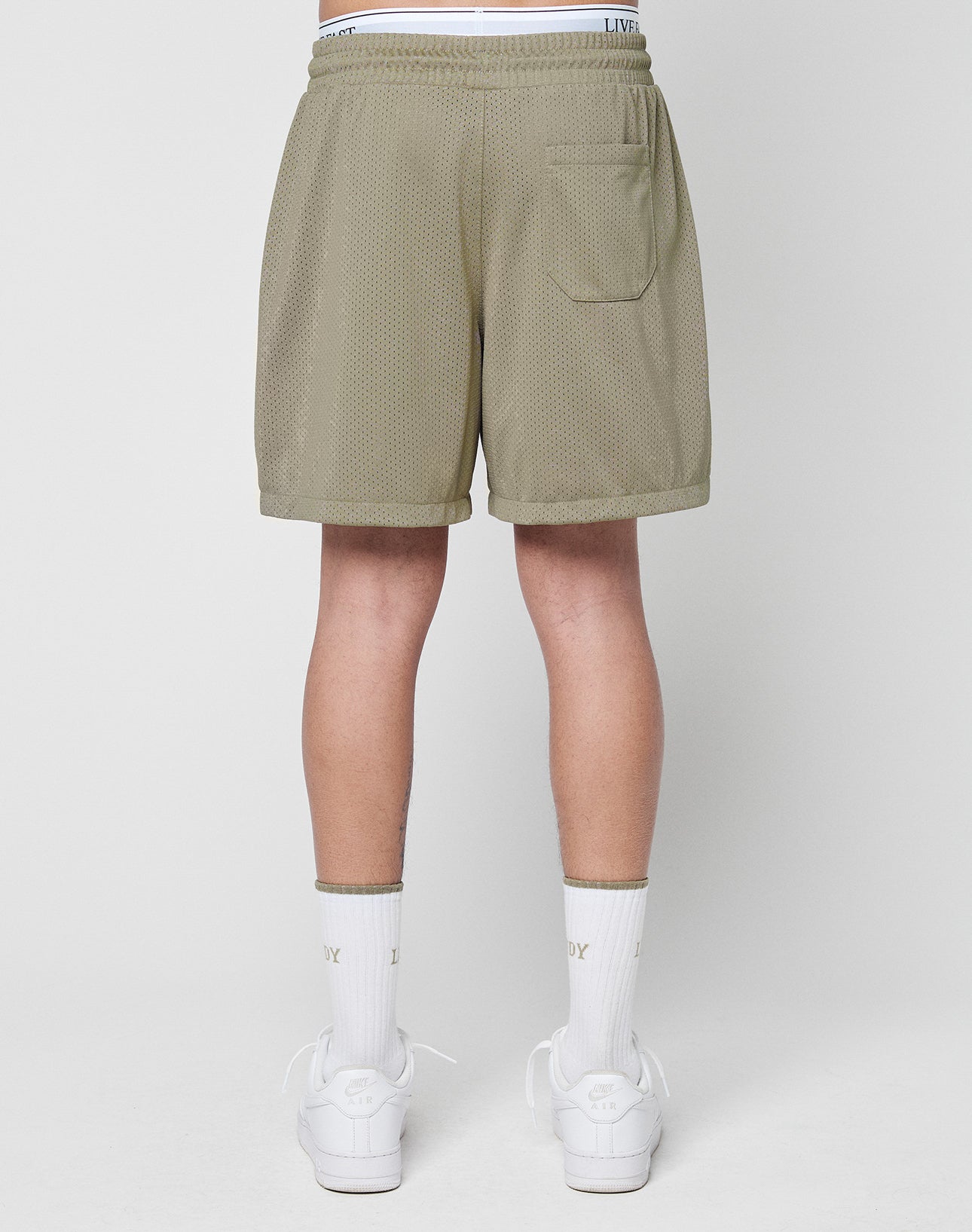 A person stands with their back to the camera, wearing LIVE FAST DIE YOUNG Basic Mesh Shorts in olive green with elastic waistband and logo patch, paired with white text socks and white sneakers, against a plain white background.
