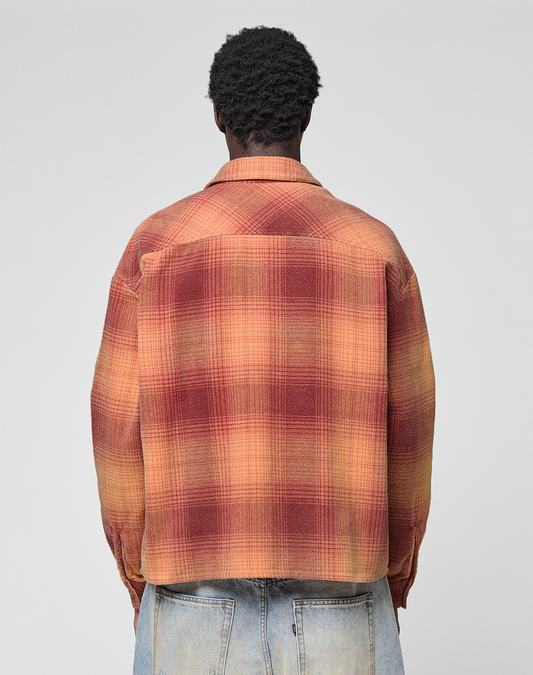 A person with short curly hair is seen from behind, wearing a loose fit red and orange LIVE FAST DIE YOUNG Vintage Flannel and light blue vintage-wash jeans against a plain light gray background.