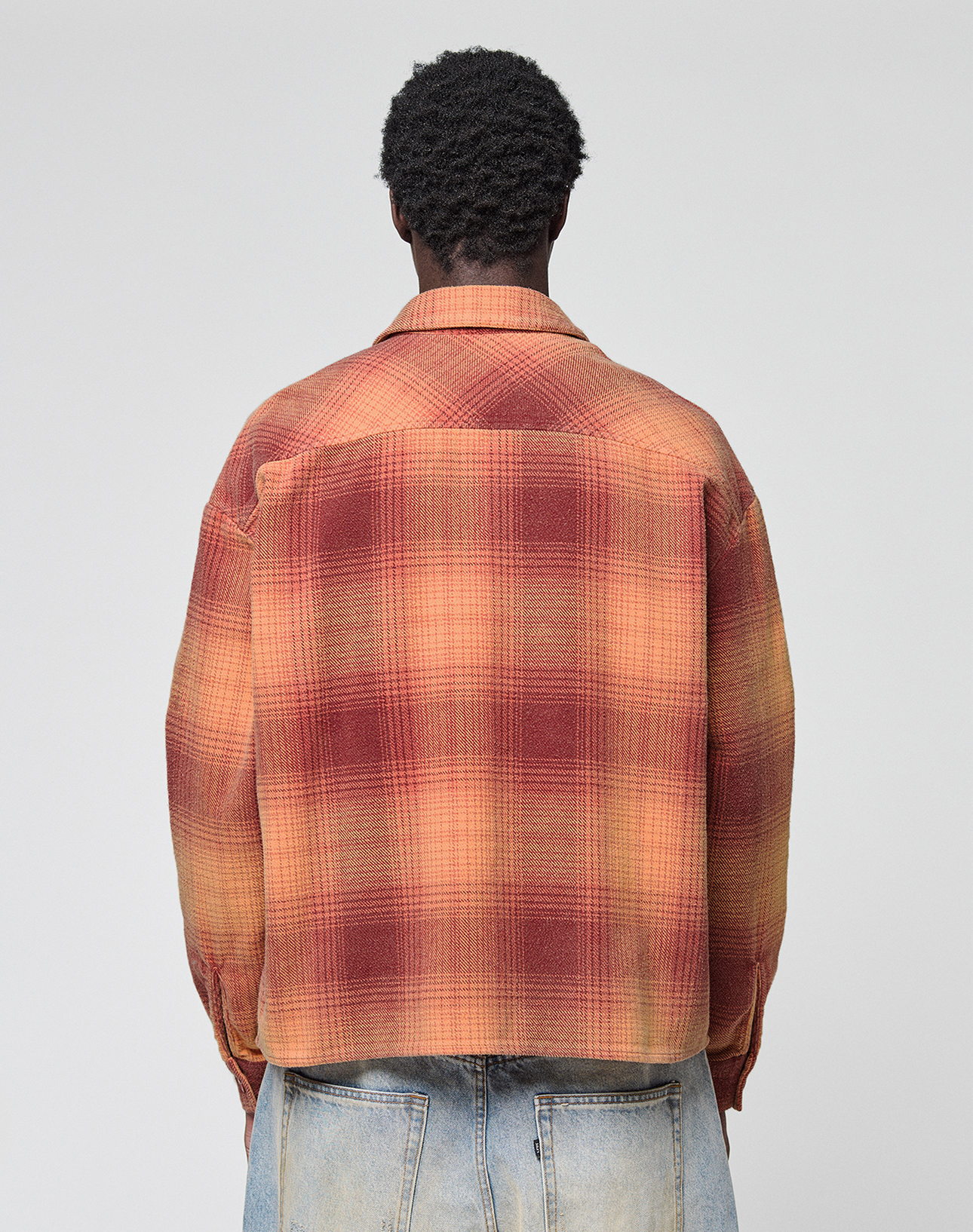 A person with short curly hair is seen from behind, wearing a loose fit red and orange LIVE FAST DIE YOUNG Vintage Flannel and light blue vintage-wash jeans against a plain light gray background.