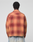 A person with short curly hair is seen from behind, wearing a loose fit red and orange LIVE FAST DIE YOUNG Vintage Flannel and light blue vintage-wash jeans against a plain light gray background.