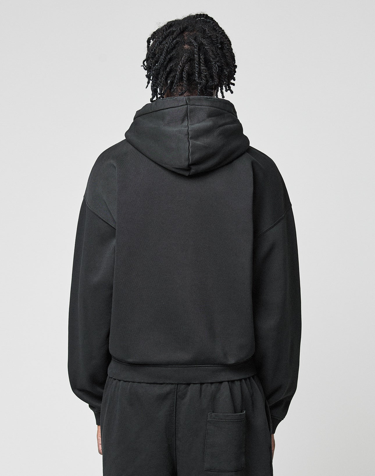 A person with short, twisted hair is seen from behind in a black LIVE FAST DIE YOUNG Basic Hooded Zip and matching black pants, standing against a plain light gray background.