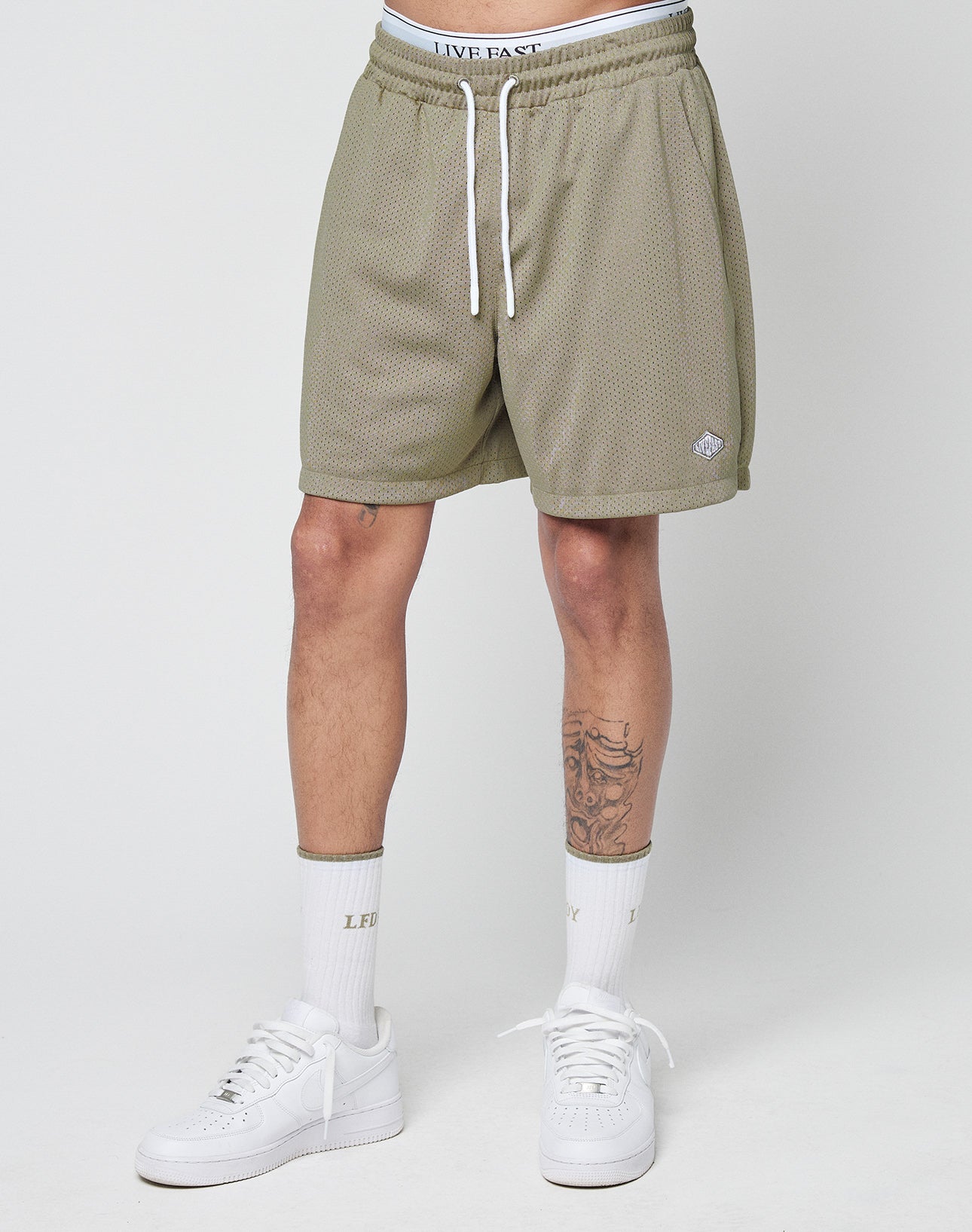 A person wears LIVE FAST DIE YOUNG Basic Mesh Shorts in olive green with a logo patch and elastic waist, white crew socks with LFT and LXY in gold, and white sneakers, standing against a plain light background—only legs and waist visible.