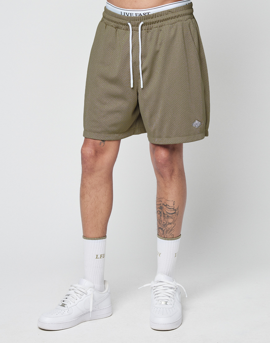 A person wears olive green Basic Mesh Shorts by LIVE FAST DIE YOUNG with a white logo patch and elastic waistband, paired with white “LIVE FAST” socks and white sneakers. Their tattooed legs stand out against a plain light background.