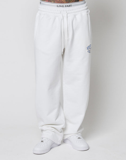 A person in LIVE FAST DIE YOUNG LFDY Radiator Sweatpants—loose-fitting, oversize, with a blue chenille patch on the left thigh and visible LIVE FAST waistband—paired with white sneakers, stands against a plain white background.