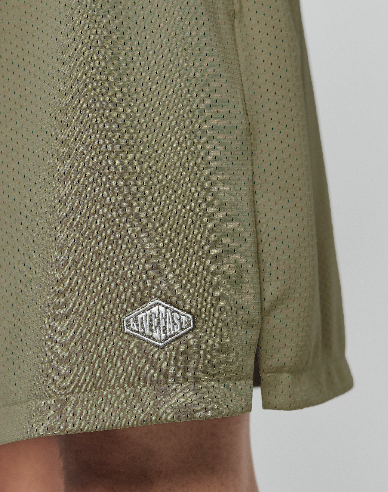 Close-up of olive green Basic Mesh Shorts by LIVE FAST DIE YOUNG, featuring a small white and gray logo patch near the hem. The athletic shorts are shown against a plain background with part of a person's lower leg visible.