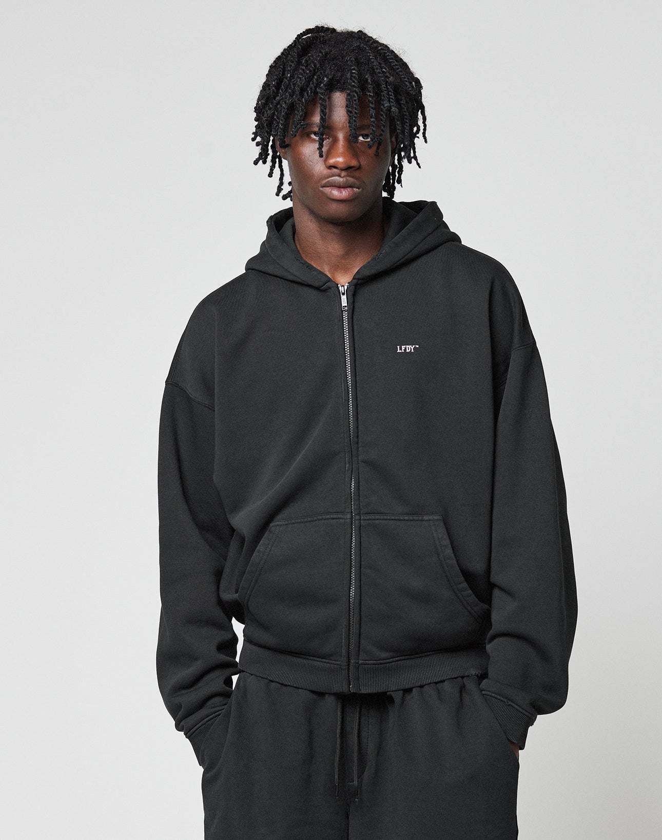 A person with short, twisted braids wears a black LIVE FAST DIE YOUNG Basic Hooded Zip and black pants, standing against a light gray background with hands in their pockets.