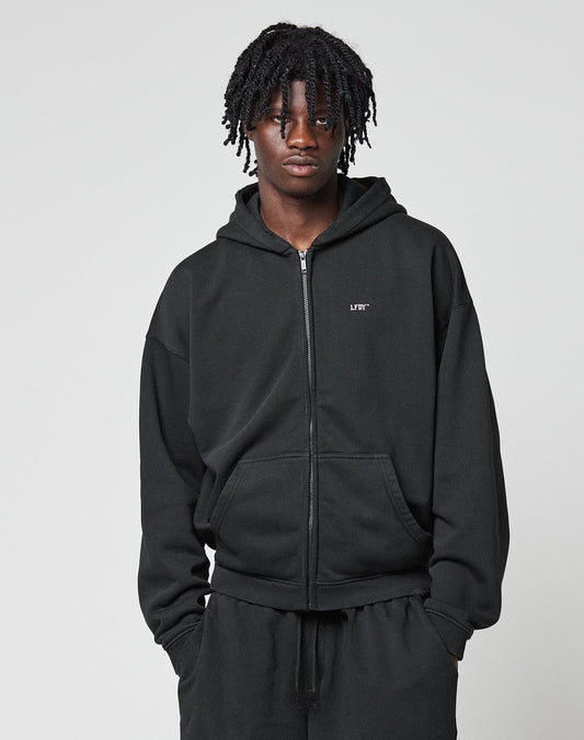A person with short, twisted braids wears a black LIVE FAST DIE YOUNG Basic Hooded Zip and black pants, standing against a light gray background with hands in their pockets.