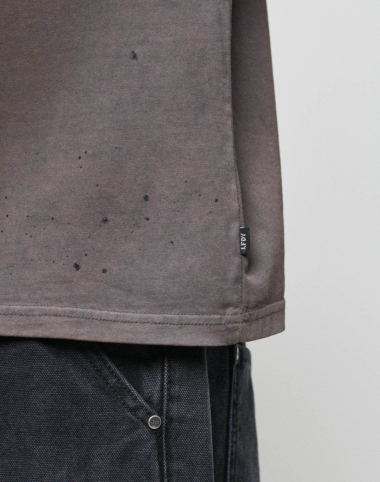 Close-up of the lower part of the LFDY Flames Muscle Shirt by LIVE FAST DIE YOUNG, a brownish-grey 100% Baumwolle tee with black specks and handgefertigtem Sprayeffekt, paired with dark denim jeans. Black fabric tag at the side seam near the hem.
