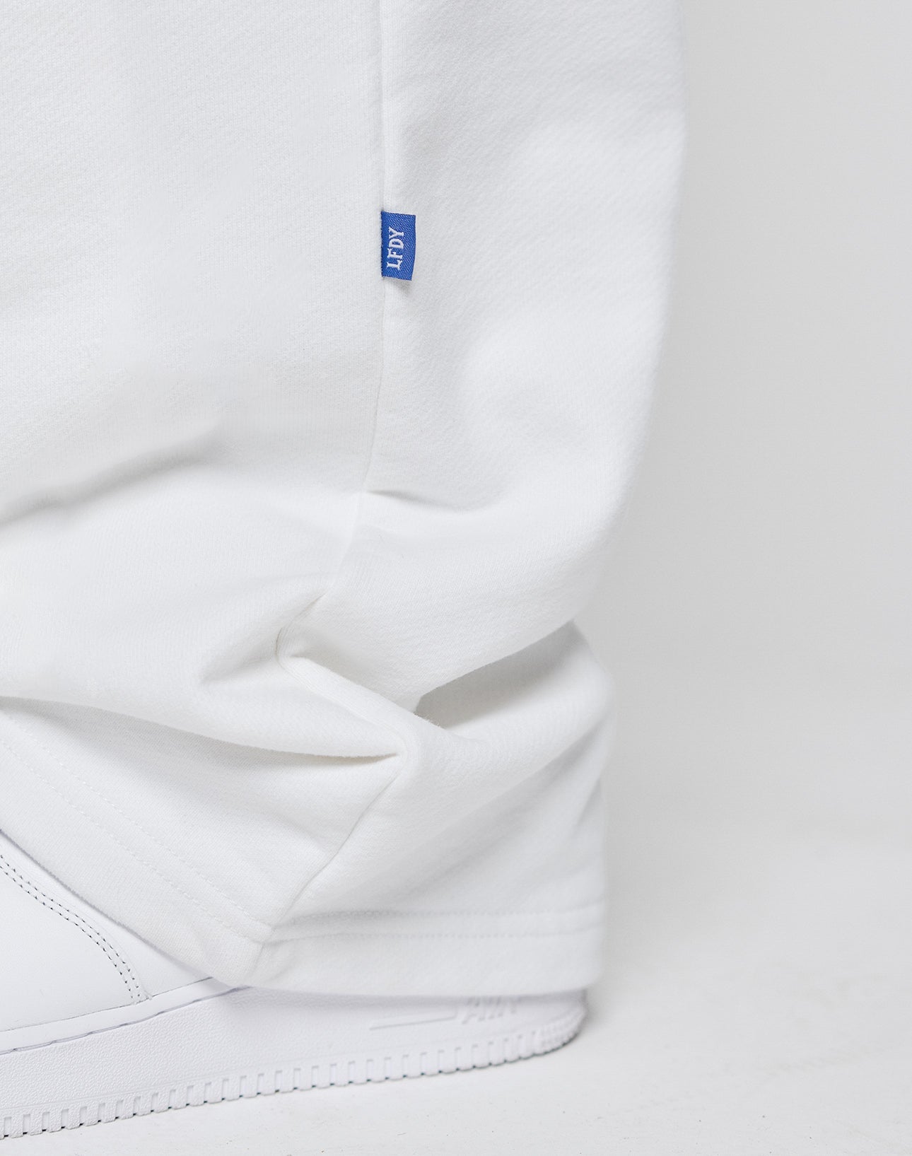 A close-up of a person wearing LIVE FAST DIE YOUNG LFDY Radiator Sweatpants in an oversize fit with a small blue seam tag and white sneakers, set against a white background.