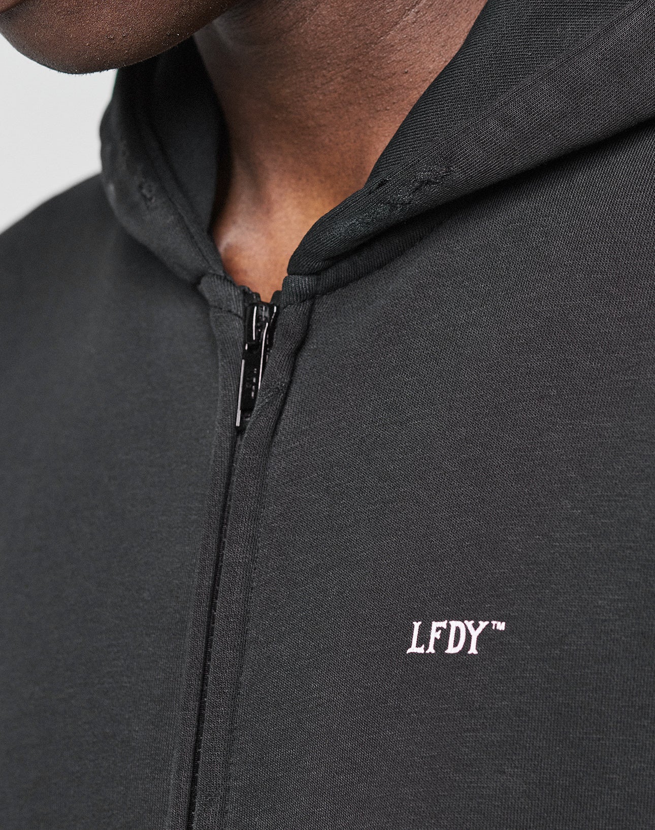Close-up of a person in a black LIVE FAST DIE YOUNG Basic Hooded Zip with the hood up and distressed Kapuze. The hoodie features a white embroidered logo on the chest, partially obscuring the persons face and neck.