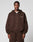 A man wears an oversize fit, dark brown LFDY Pleated Hooded Zip by LIVE FAST DIE YOUNG with LIFE IS PART text on the left chest and matching sweatpants, standing against a plain light gray background.