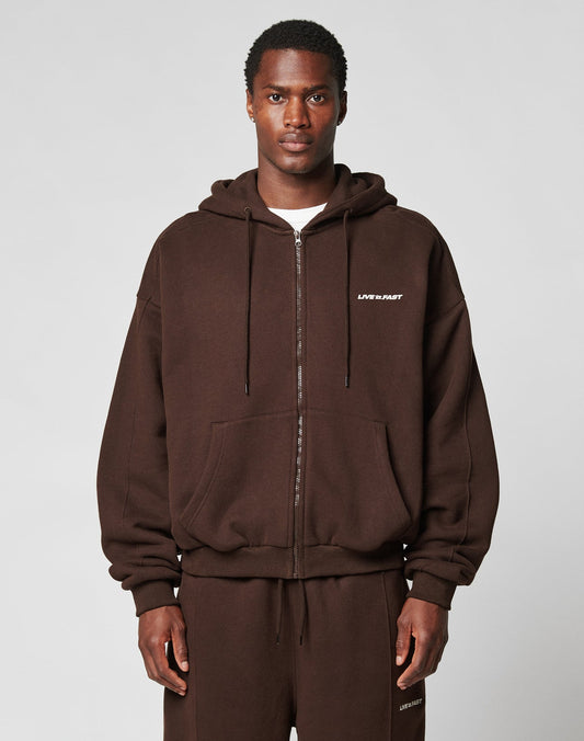 A man wears an oversize fit, dark brown LFDY Pleated Hooded Zip by LIVE FAST DIE YOUNG with LIFE IS PART text on the left chest and matching sweatpants, standing against a plain light gray background. | model
