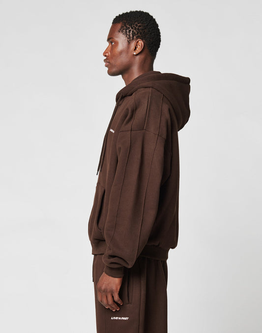 A man stands in profile against a plain background, wearing an oversize fit LFDY Pleated Hooded Zip by LIVE FAST DIE YOUNG with the hood down and matching brown sweatpants.