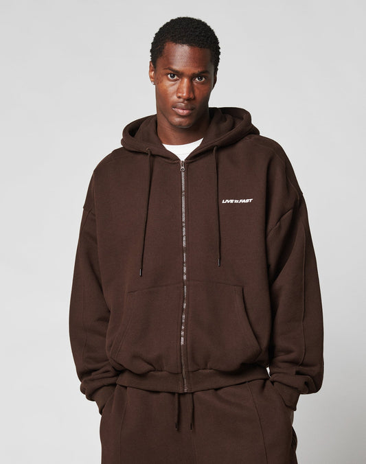 A man stands against a plain light background, hands in his pockets, wearing the LFDY Pleated Hooded Zip by LIVE FAST DIE YOUNG—an oversize fit brown zip-up hoodie aus 100% Baumwolle with a small white LIVE FAST logo and Kapuze mit Kordel.