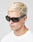 A man with short, bleached blond hair wears LIVE FAST DIE YOUNGs Live Fast Curved Shades—black rectangular Mazzuchelli Acetate sunglasses with 400 UV protection and LIVE FAST on the side—paired with a plain white shirt against a light background.
