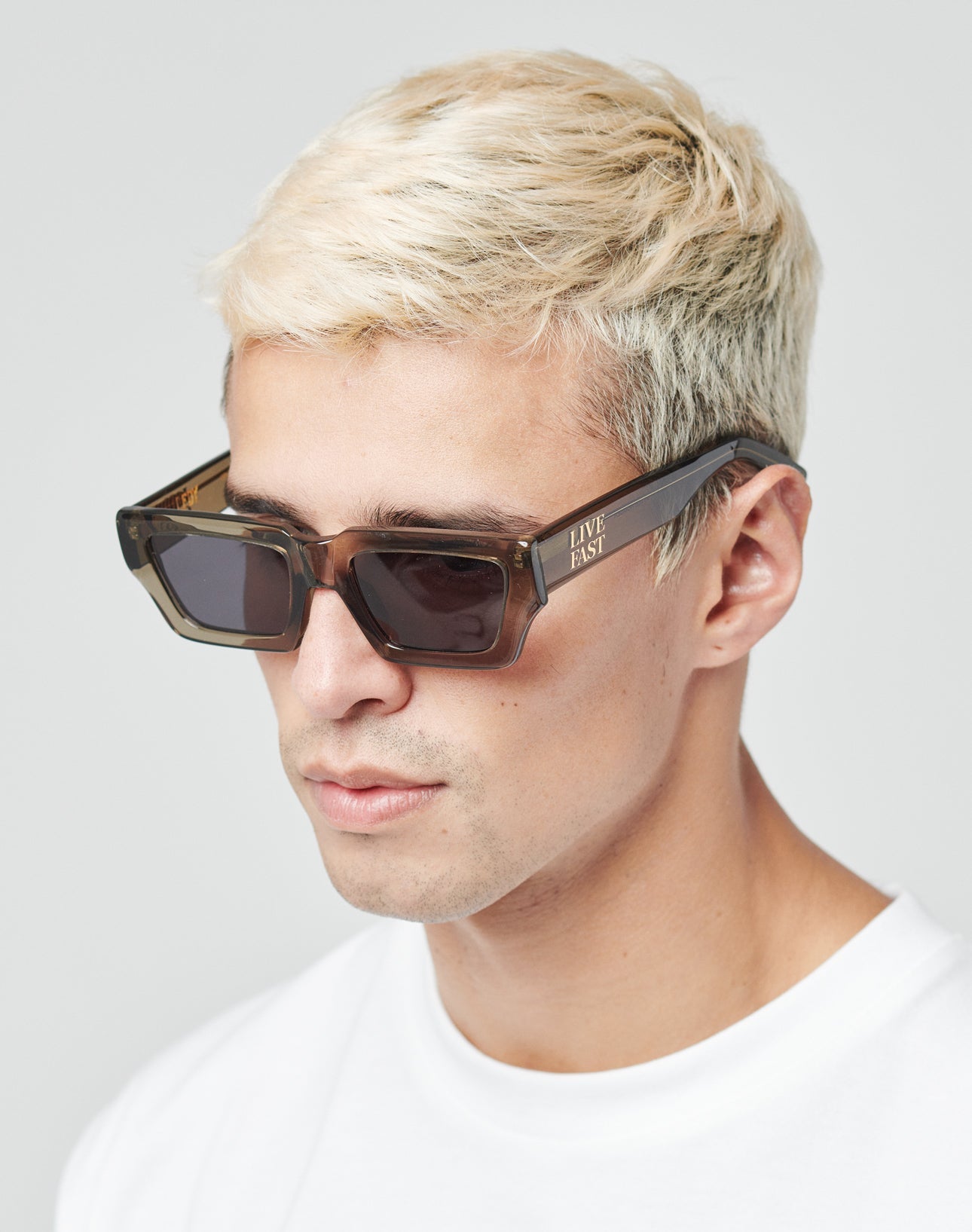 A young man with short platinum blonde hair wears LIVE FAST DIE YOUNGs Live Fast Sunglasses—angular, thick-rimmed Mazzuchelli Acetate frames with LIVE FAST on the side and a gold logo—paired with a white T-shirt against a plain background.
