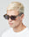 A young man with short platinum blonde hair wears LIVE FAST DIE YOUNGs Live Fast Sunglasses—angular, thick-rimmed Mazzuchelli Acetate frames with LIVE FAST on the side and a gold logo—paired with a white T-shirt against a plain background.