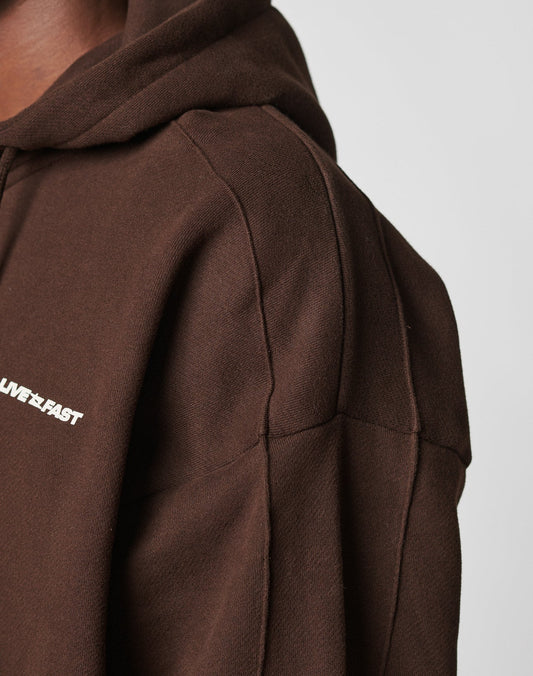 Close-up of a person in the LFDY Pleated Hooded Zip by LIVE FAST DIE YOUNG, featuring brown fabric, an Oversize Fit, partial white LIVE FAST text on the chest, and visible detailed stitching on the shoulder and upper arm.