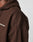 Close-up of a person in the LFDY Pleated Hooded Zip by LIVE FAST DIE YOUNG, featuring brown fabric, an Oversize Fit, partial white LIVE FAST text on the chest, and visible detailed stitching on the shoulder and upper arm.
