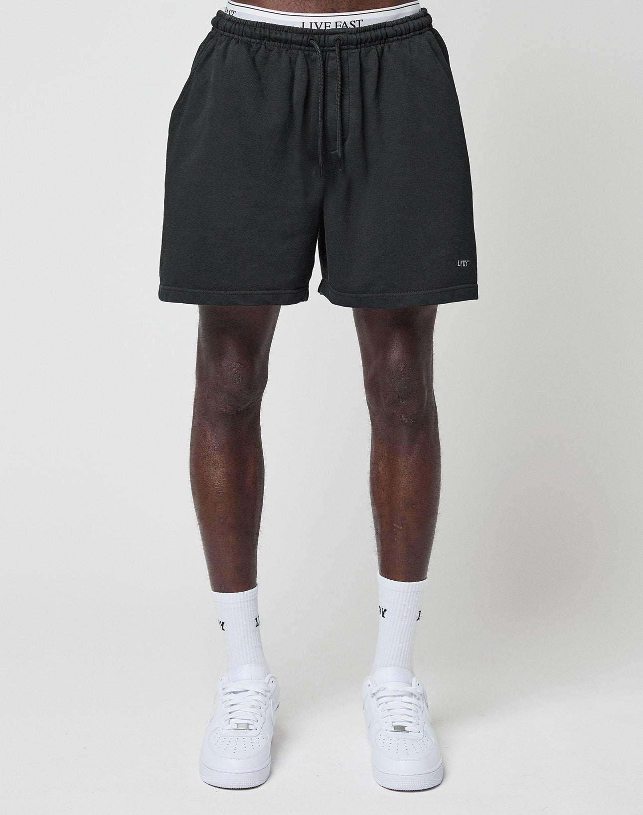 A person wears LIVE FAST DIE YOUNG Basic Sweat Shorts with LIVE FAST on the elastic waistband, paired with white socks and sneakers, standing against a plain light gray background.