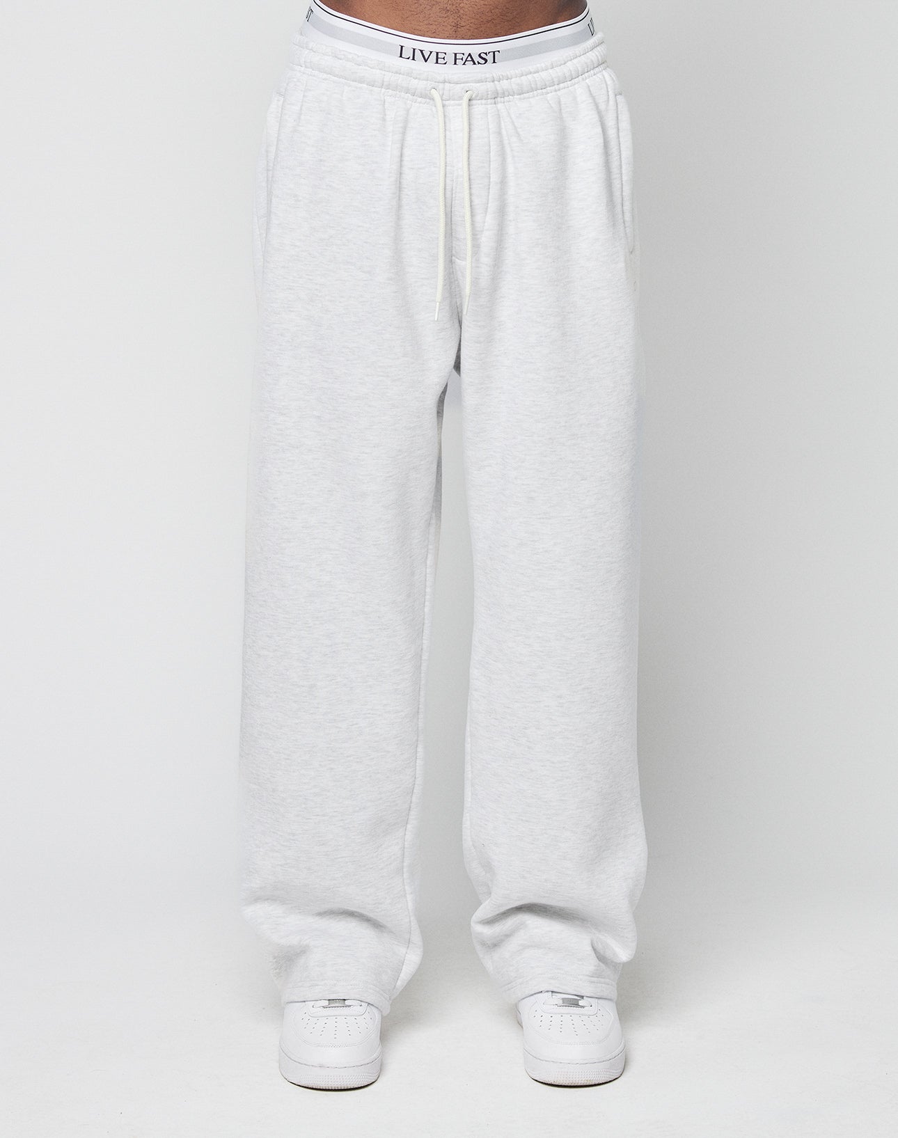 A person wears light gray Basic Wide Jogger sweatpants by LIVE FAST DIE YOUNG with an elastic waistband and drawstring, white sneakers, and a visible white waistband featuring LIVE FAST, standing against a plain white background.