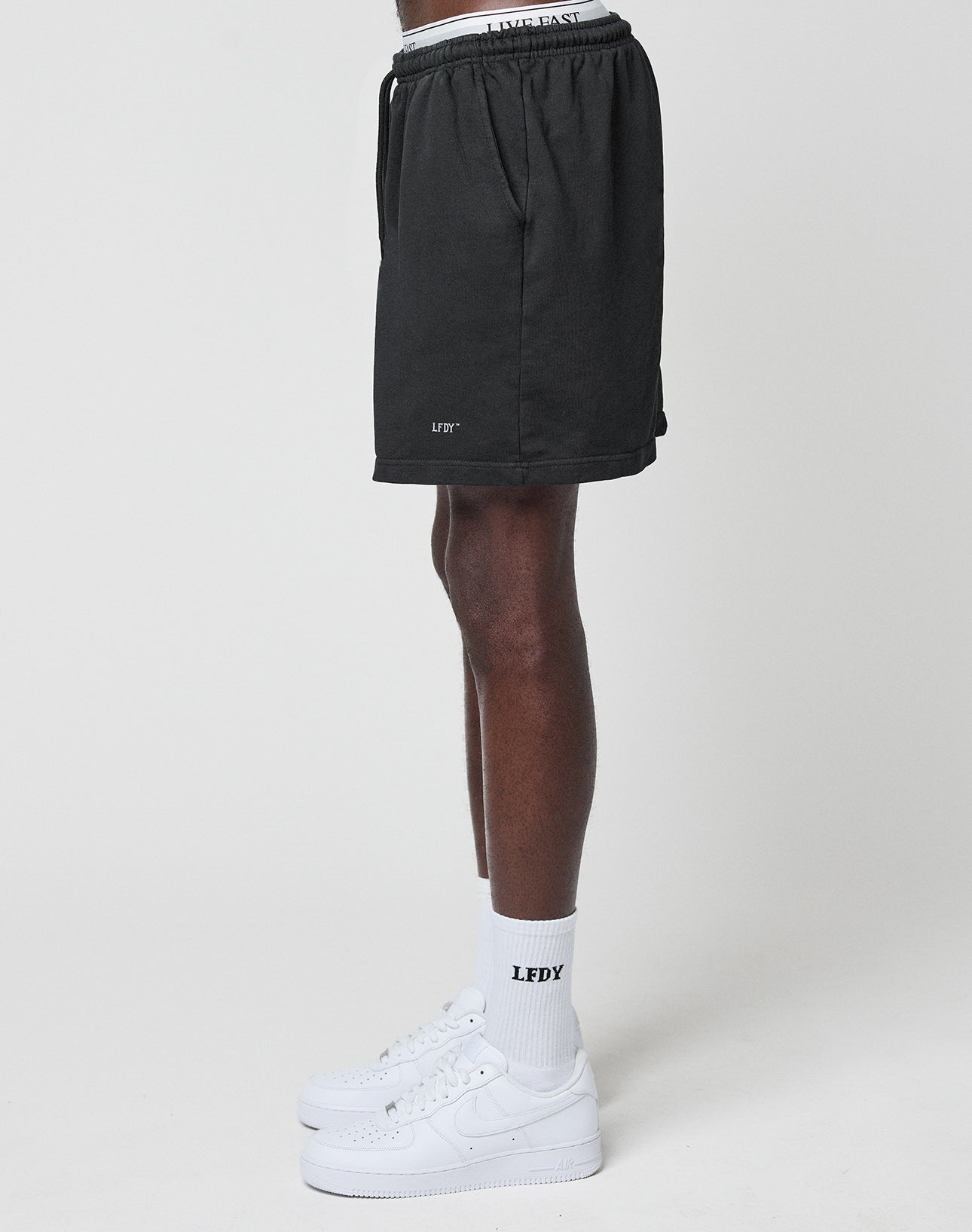 A person stands in profile against a plain white background, wearing LIVE FAST DIE YOUNG Basic Sweat Shorts in black with an elastic waistband, LFDY Logo Print white socks, and white sneakers. Only the lower half of their body is visible.